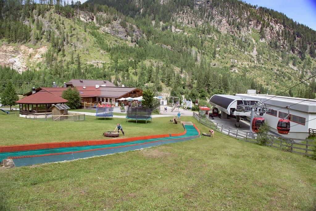 A leisure area in the mountains with a toboggan run and a restaurant. In the background, a cable car and green hills can be seen.