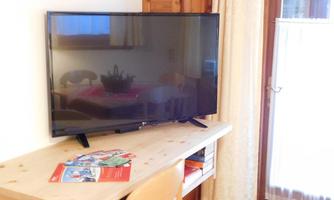 A television is standing on a wooden shelf. In the background, a table and a window with curtains are visible.