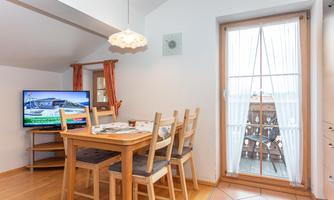 A cozy dining area with a wooden table and chairs. In the background, there is a television and a door leading to a balcony.