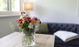 A colorful bouquet in a glass vase is sitting on a wooden table. In the background, a cozy sofa and a lamp can be seen.