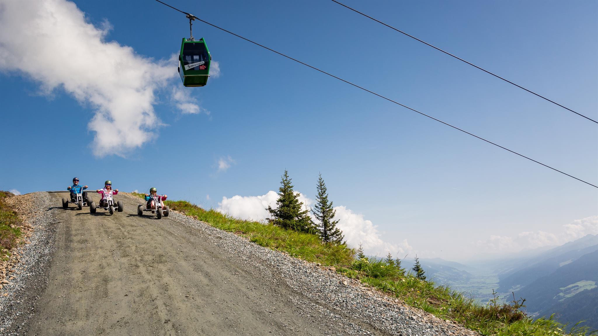 A group of people is racing down a mountainside in go-karts. In the background, there is a cable car and a clear blue sky.