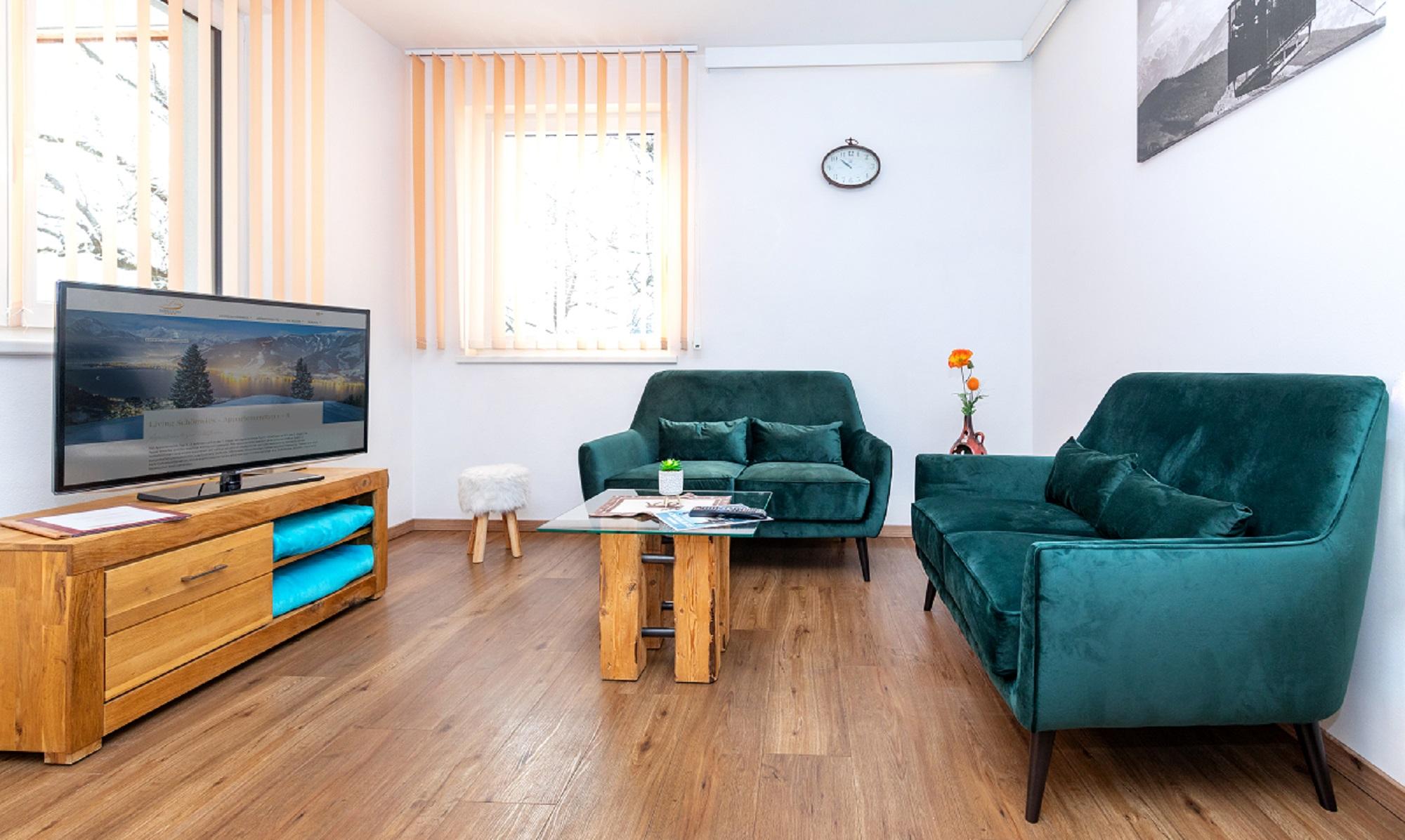 A modern living room with two green sofas and a wooden coffee table. A television stands on a piece of furniture, and there is a flower on the table.
