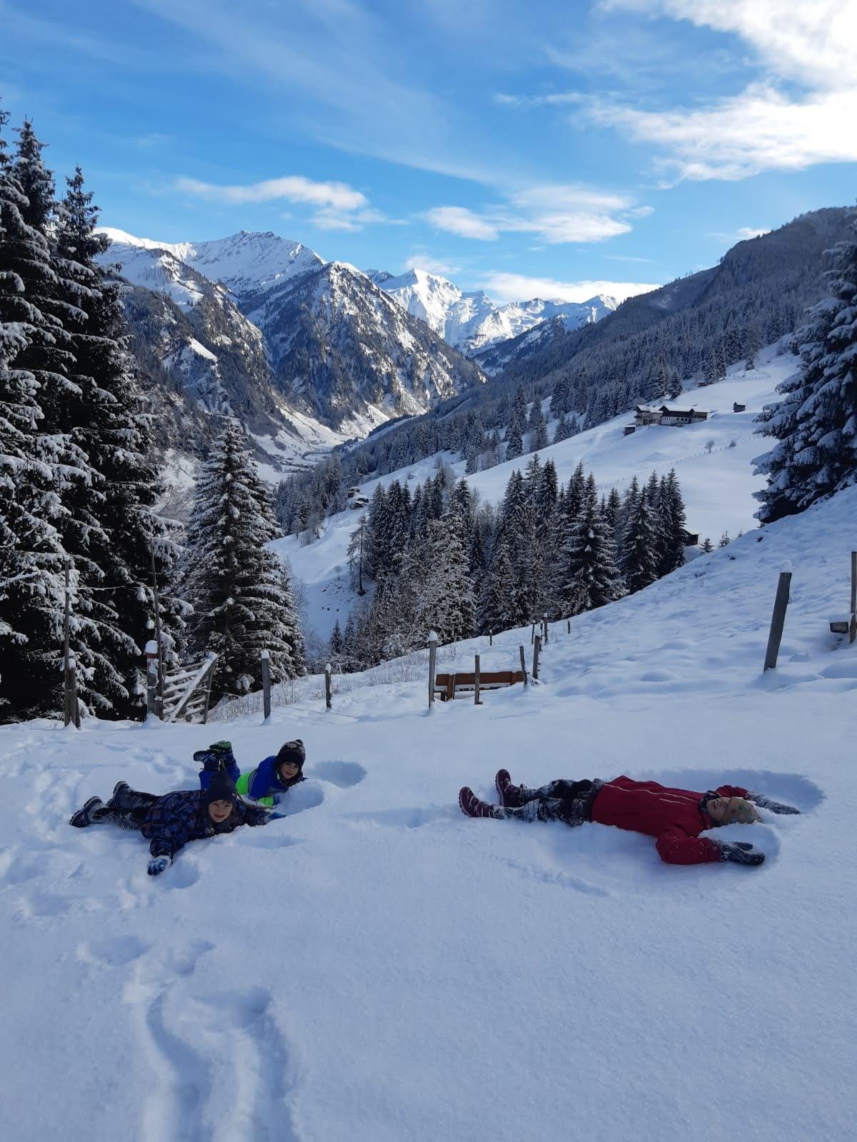 Two children are lying in the snow, enjoying the winter landscape. In the background, snow-covered mountains and pine trees can be seen.