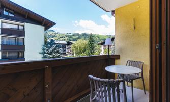 A balcony with a small table and two chairs. In the background, there are houses and green hills.