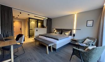 A modern hotel room with a large bed, a cozy armchair, and a table. The room is bright and stylishly decorated, with a view of a bathroom.