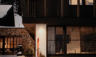 A modern house in the snow with large windows and rustic wood design. Skis stand in front of the door, and the light shines warmly into the darkness.
