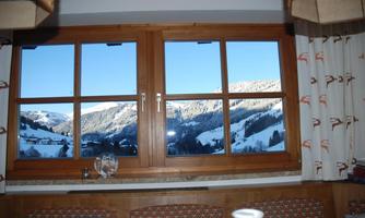 A window with a view of snowy mountains and a clear blue sky. In the foreground, cozy furniture and a coffee cup are visible.