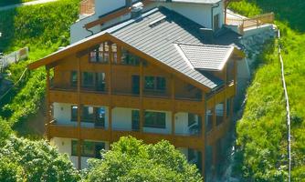 A modern wooden house surrounded by green nature. It has several windows and balconies that offer a view of the surroundings.