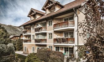 A modern residential building with several balconies and pretty plants in the foreground. The surroundings are green and surrounded by mountains.