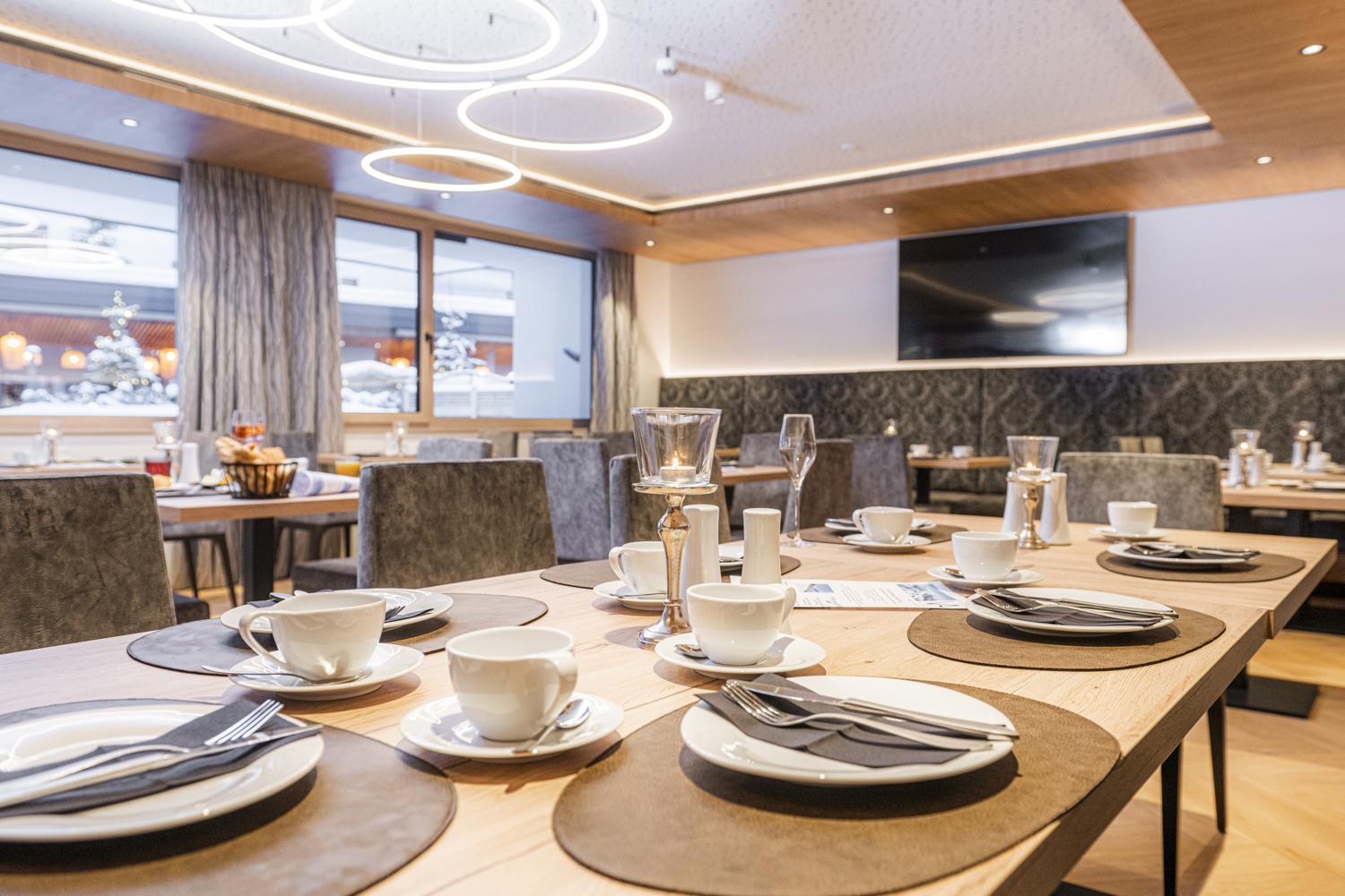 An elegant restaurant with modern design. The tables are set with plates and cups, and large windows offer a view of the surroundings.
