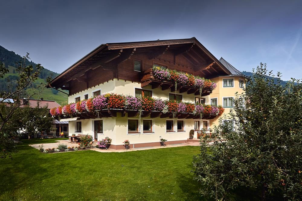 A picturesque building with balconies adorned with colorful flowers. Above it stretches a green meadow and surrounding mountains.