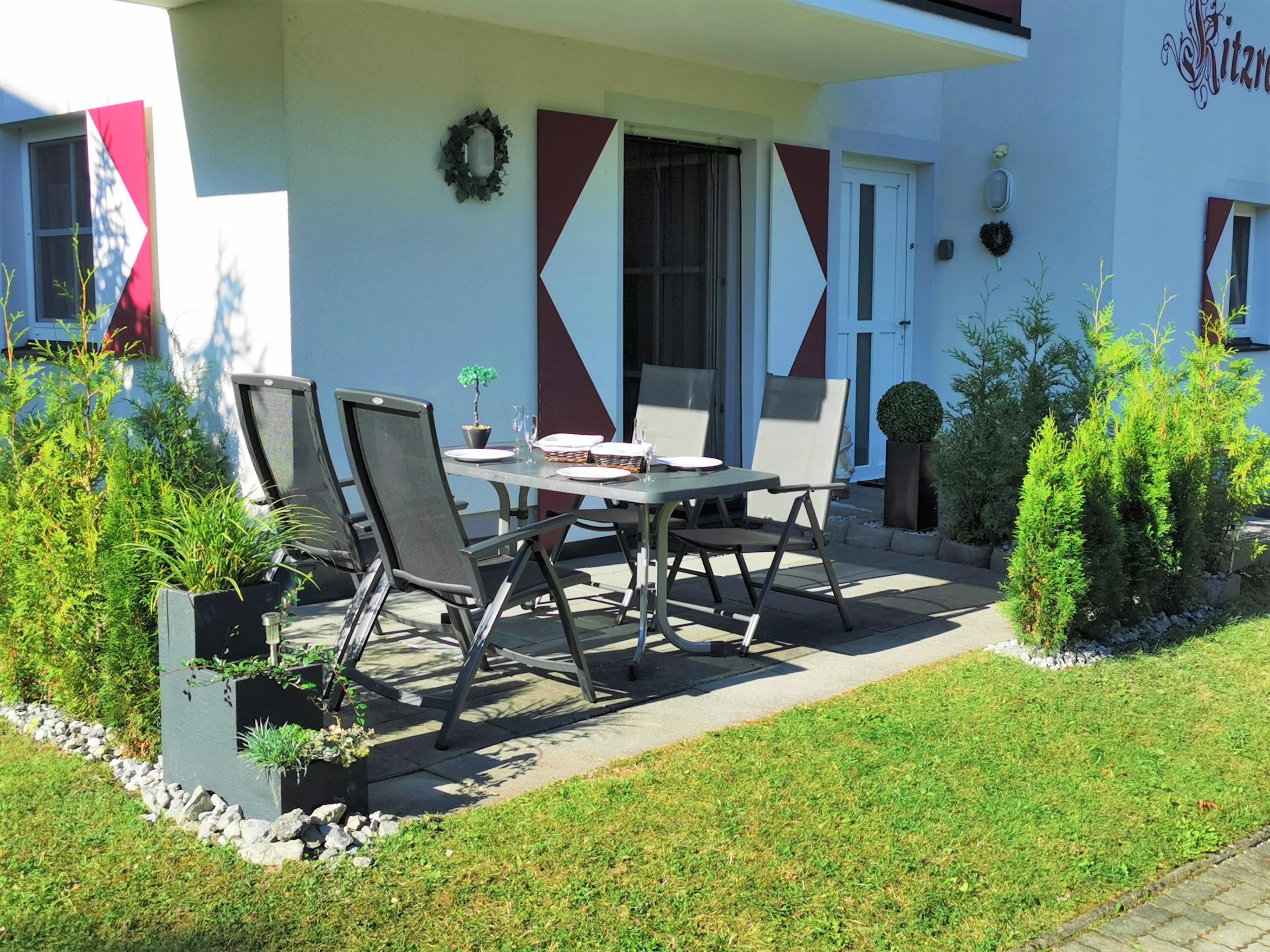 A beautiful outdoor area with a table and four chairs. Surrounded by green plants and a well-maintained lawn.
