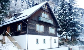 A cozy wooden house in the snow, surrounded by coniferous trees. The facade is traditional and welcoming.