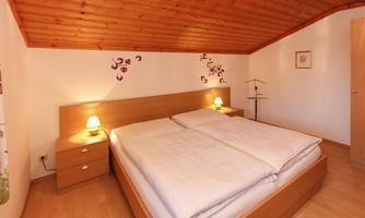 A cozy bedroom with a double bed and warm wooden decor. The walls are adorned with subtle wall tattoos, and there are nightstands with lamps.