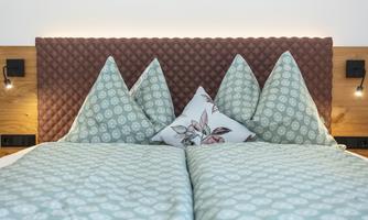 A modern bedroom with a padded headboard and colorful cushions. The bedding is in soft colors and features a subtle pattern.