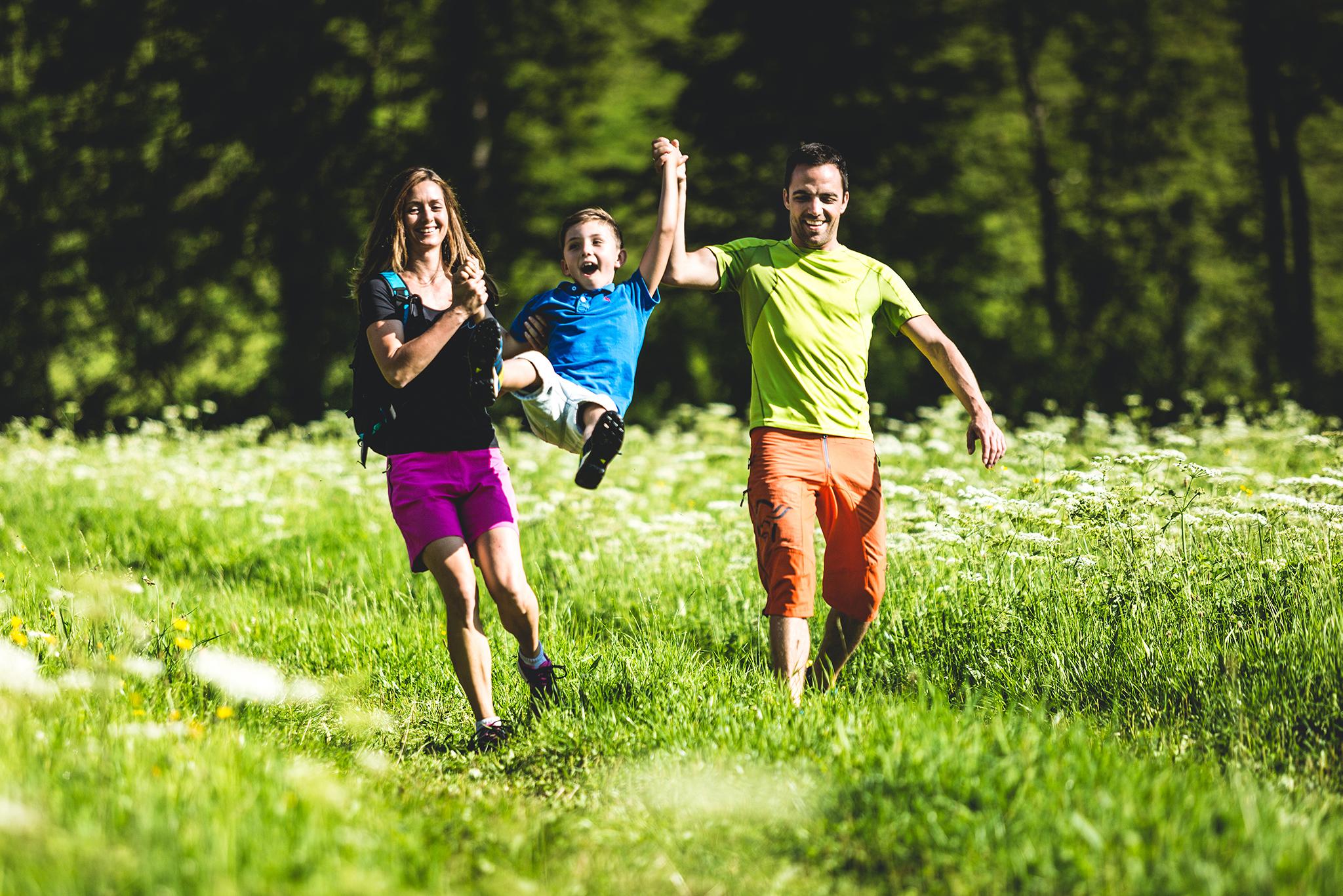 A family is happily running through a green meadow. The boy is being lifted up by his father, while the mother beams next to them.