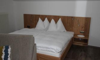 A modern bedroom with a double bed and three pillows. The walls are light, and there is a cozy seating area.