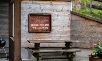 A wooden sign with the inscription "Mountain Panorama Fam Abreger" hangs on a wall. In front of the sign stands a wooden table with benches.