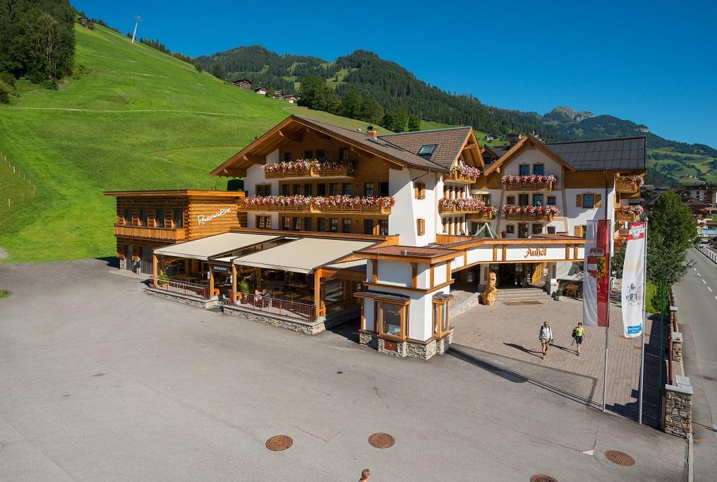 A cozy hotel in alpine style, surrounded by green hills. The sun is shining on the terrace and the building.
