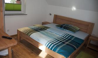 A simple bedroom with a wooden bed and a colorful duvet. There is a desk and a window with green blinds.