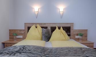 A cozy bedroom with a double bed and yellow cushions. There are modern wall lamps on the walls and plants on the nightstands.