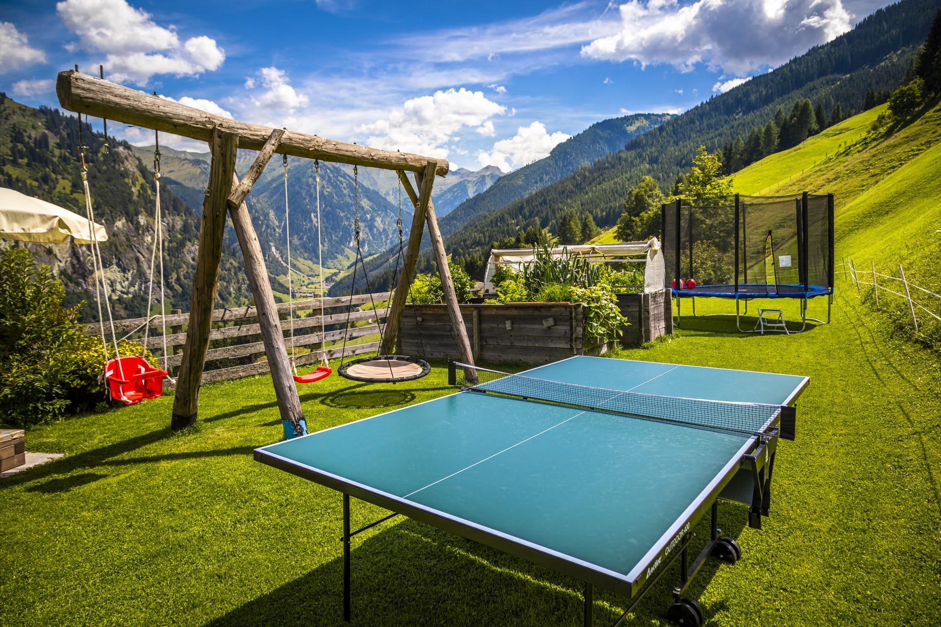 A beautiful garden with a table tennis court and a swing. In the background, green mountains stretch under a blue sky.