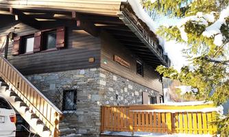 A cozy chalet with a wooden facade and stone details. It is surrounded by snow and has an inviting wooden terrace.