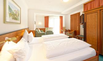 A cozy hotel room with two beds and large windows. In the background, a seating area and a desk are visible.