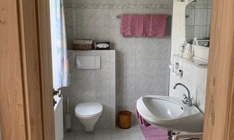 A small, bright bathroom with white tiles. There is a sink, a toilet, and pink towels.