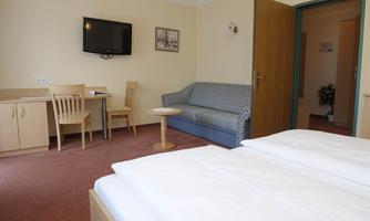 A simple hotel room with two beds, a sofa, and a small table. A television is mounted on the wall, and there is a table with chairs.