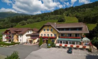 A picturesque hotel with colorful flower boxes and a beautiful mountain landscape in the background. The building has an inviting terrace and a parking lot.