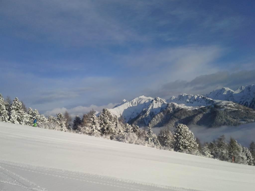 A snowy landscape with high mountains in the background. The sky is clear and blue, creating a serene winter atmosphere.