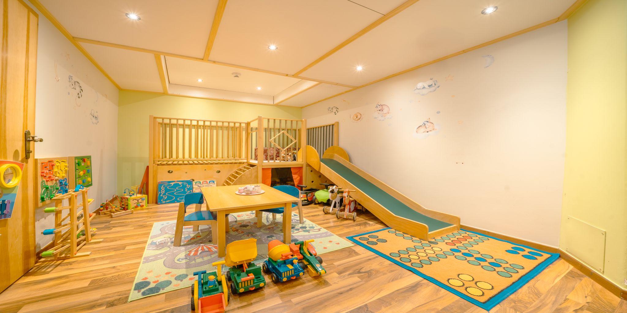A colorful playroom with a slide, a play table, and various toys. The walls are bright and cheerful.