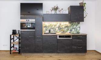 A modern kitchen with dark furniture and a wooden work area. In the background, there is a decorative wall with plant motifs.