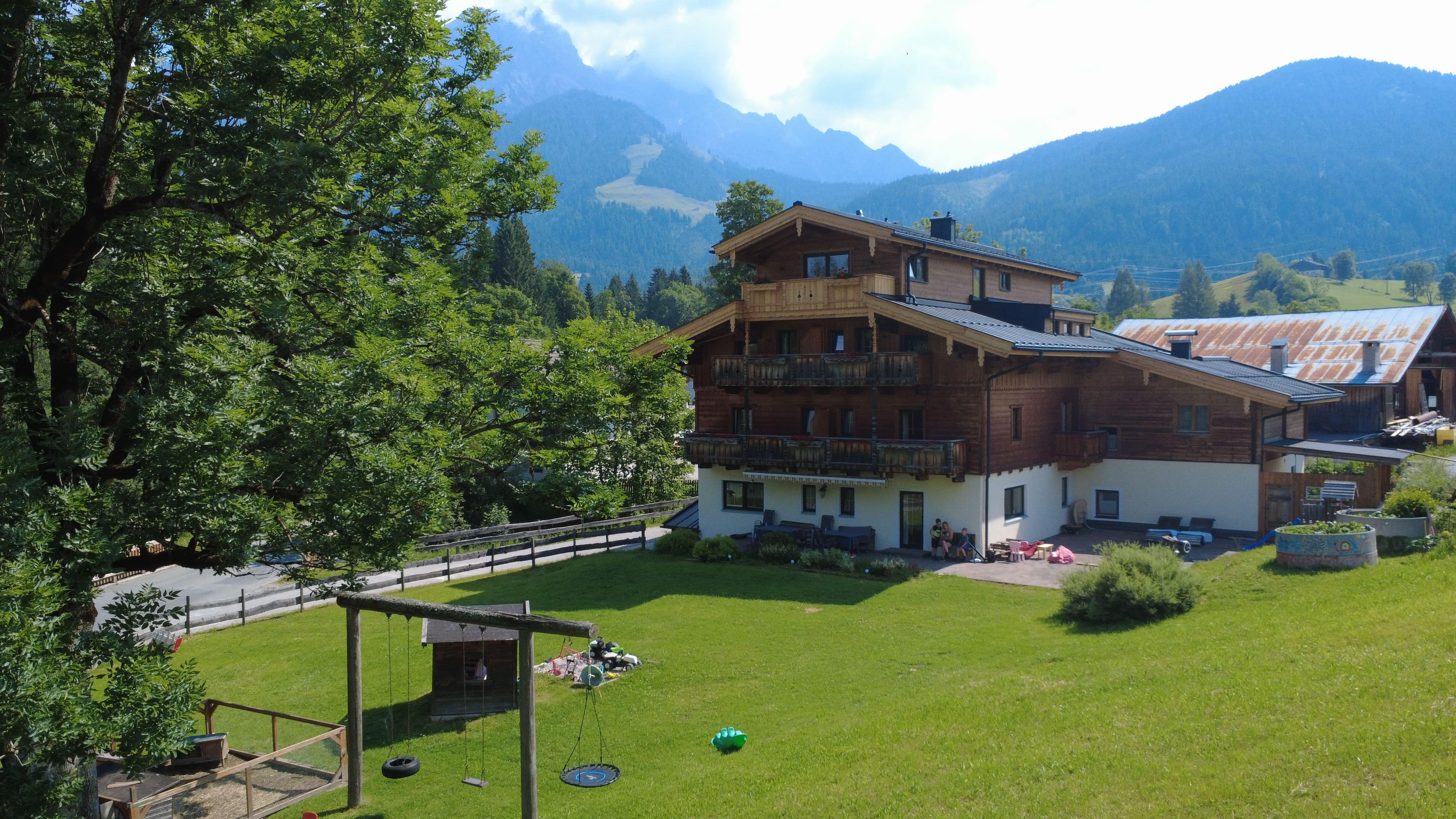 A traditional house amidst a green landscape, surrounded by mountains. The garden is well-kept and has plenty of space to relax.