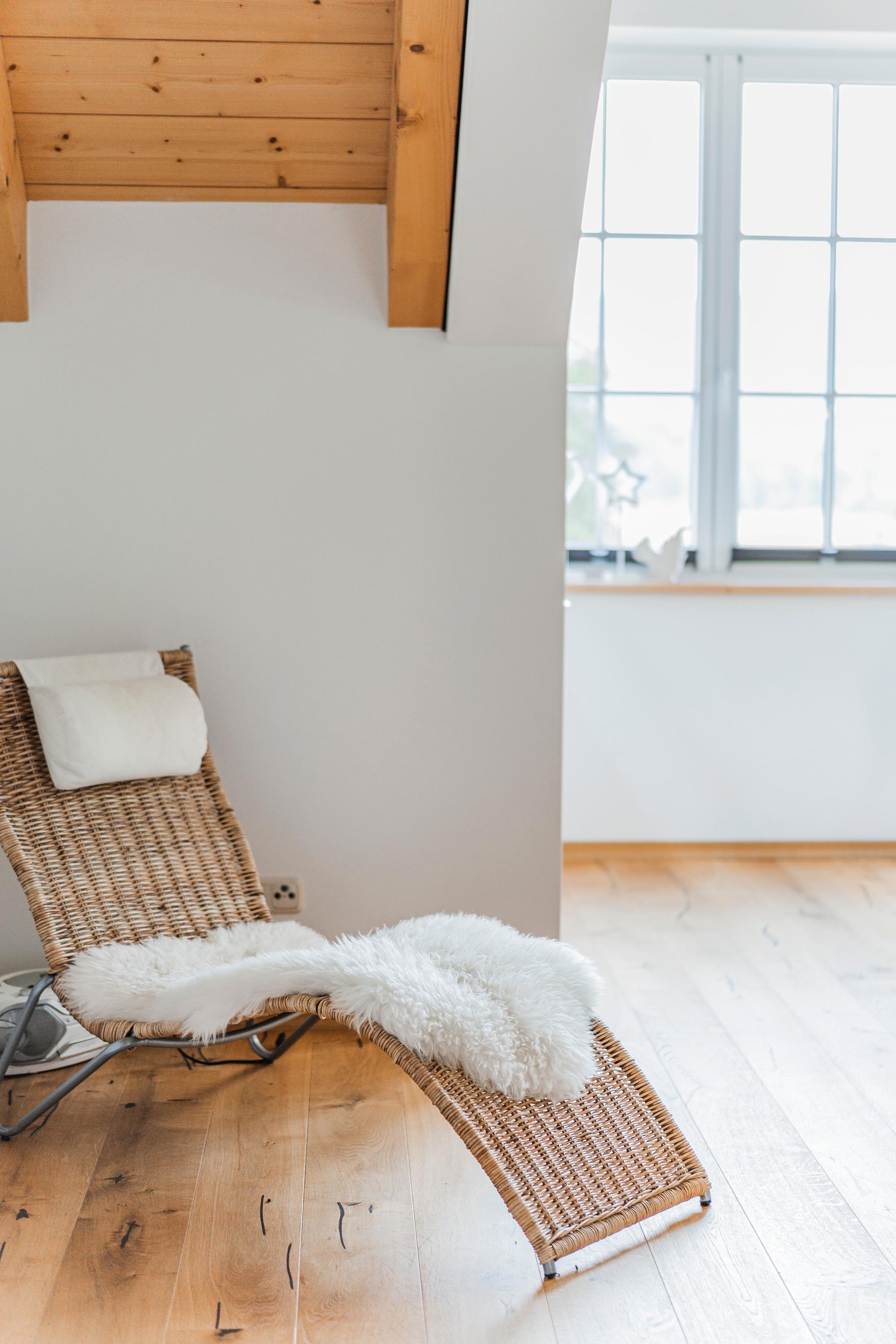 A modern wooden lounge chair is in a bright room with large windows. The floor is made of light wood, and the room feels friendly and inviting.