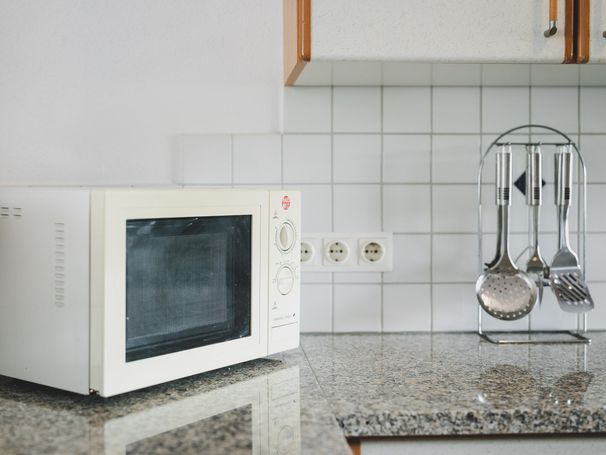 A modern microwave stands on a granite countertop in a kitchen. In the background, cooking utensils are visible on a holder and a tiled wall.