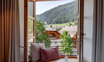 A window view of a picturesque mountain landscape. In the foreground, there are cozy cushions and a small table.