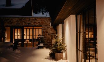 A snow-covered outdoor area with a warmly lit house. The cozy atmosphere evokes a winter evening.