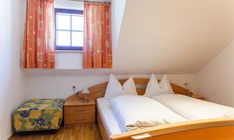 A cozy bedroom with a double bed and bright pillows. Orange curtains hang in front of a window, and a patterned bench stands in the corner.