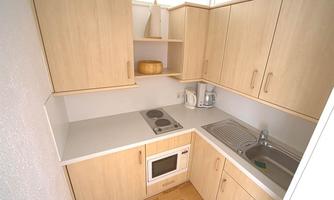 A small modern kitchen with light wood cabinets. It features a stove, a sink, and a microwave.