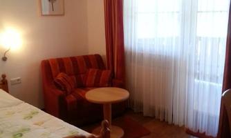 A cozy room with a single bed, a red armchair, and a table. Large windows let in plenty of daylight.
