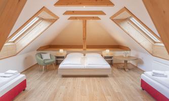 A bright and modern attic room with wooden beams. It is equipped with a double bed, two single beds, and stylish furniture.