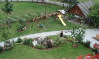 A playground with a slide, a swing, and a small playhouse is seen in a green environment. The scene is surrounded by trees and garden plants.