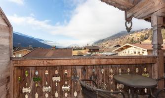 A balcony with a table and chairs offers a view of a picturesque mountain landscape. The sky is partly cloudy and the surroundings feel calm and inviting.
