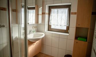 A bright bathroom with a sink, a mirror, and a shower cubicle. A window with a white curtain provides natural light.