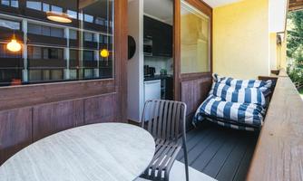 A cozy balcony with a round table and a comfortable, striped armchair. The walls are made of wood, and there are large windows that let in plenty of light.