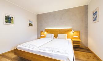 A modern bedroom with a double bed and soft wooden tones. Pictures hang on the wall, and a warm white lighting creates a cozy atmosphere.