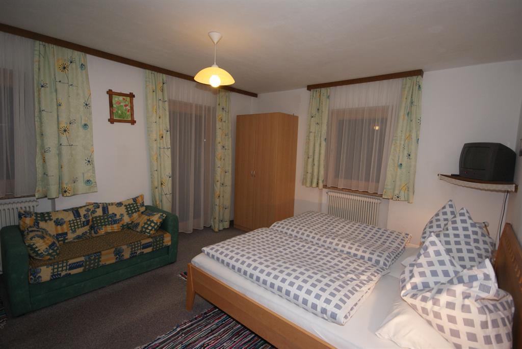 A simple bedroom with a double bed and a green sofa. The walls are light and decorated with floral-patterned curtains.
