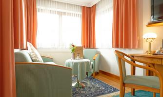 A cozy living area with two armchairs and a small table. The windows are dressed with light curtains and the room is friendly and inviting.
